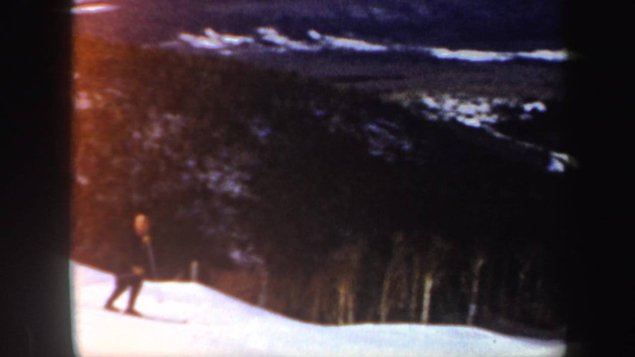 Skier in dark clothing on a snowy slope with trees and distant hills behind.