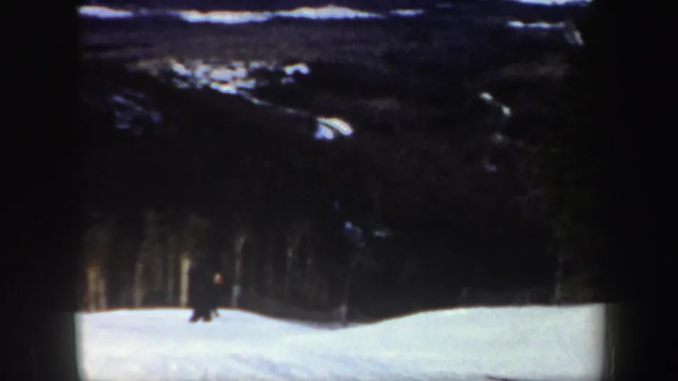 A small dark figure skis along the lower edge of the snowy hill, with soft-focus trees and distant ridges behind.