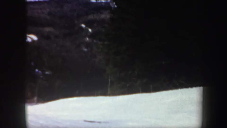 The skier is mostly at the left edge as the camera holds on the snowy slope, wooded hillside and dark ridgeline beyond.
