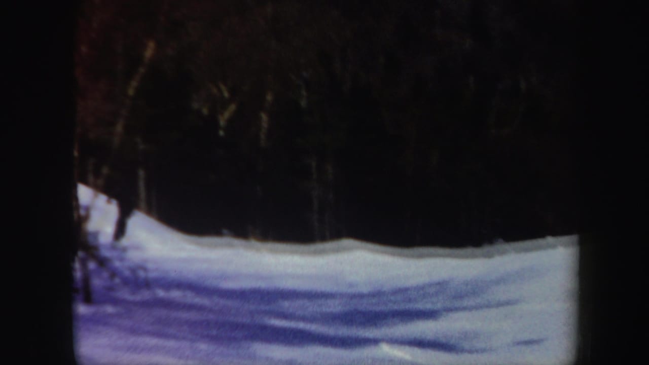 1962 Vermont winter hillside on 8mm film, with snow, tree shadows, and dark woods at the top of the frame.