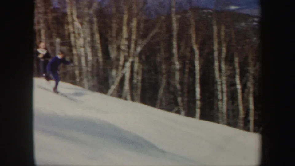 1962 Vermont 8mm frame of a skier in blue moving down a snowy forest slope with birch trees behind.