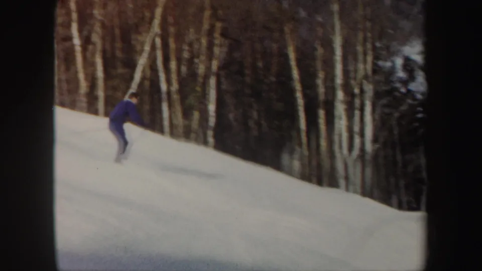 The skier in blue moves farther across the Vermont snow slope, backed by dense bare trees and evergreens.