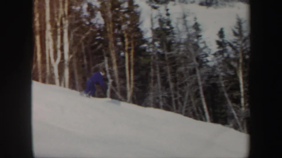 A blue-clad skier crouches low on the snowy Vermont slope near birch trunks and darker evergreens.