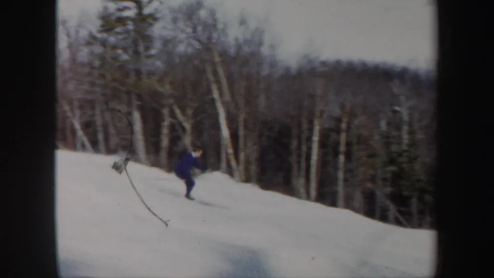 1962 Vermont home-movie frame showing a skier in blue on a snow-covered hillside bordered by birch trees and evergreens.