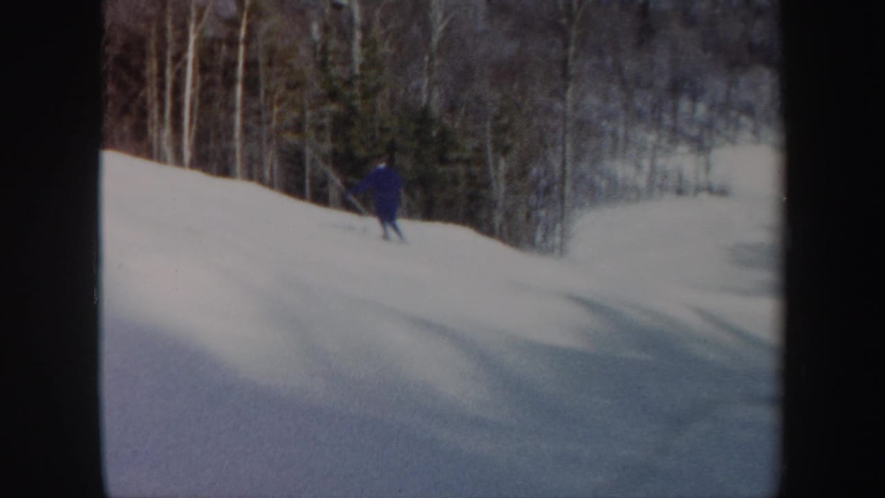 A distant skier in blue moves down a Vermont snow slope, with leafless trees and evergreens in the background.