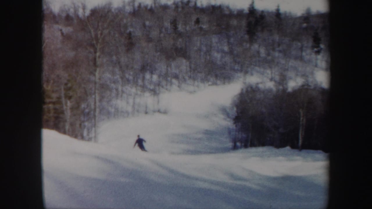 The skier continues downhill on a wooded Vermont ski slope, small in frame and surrounded by pale snow and winter trees.