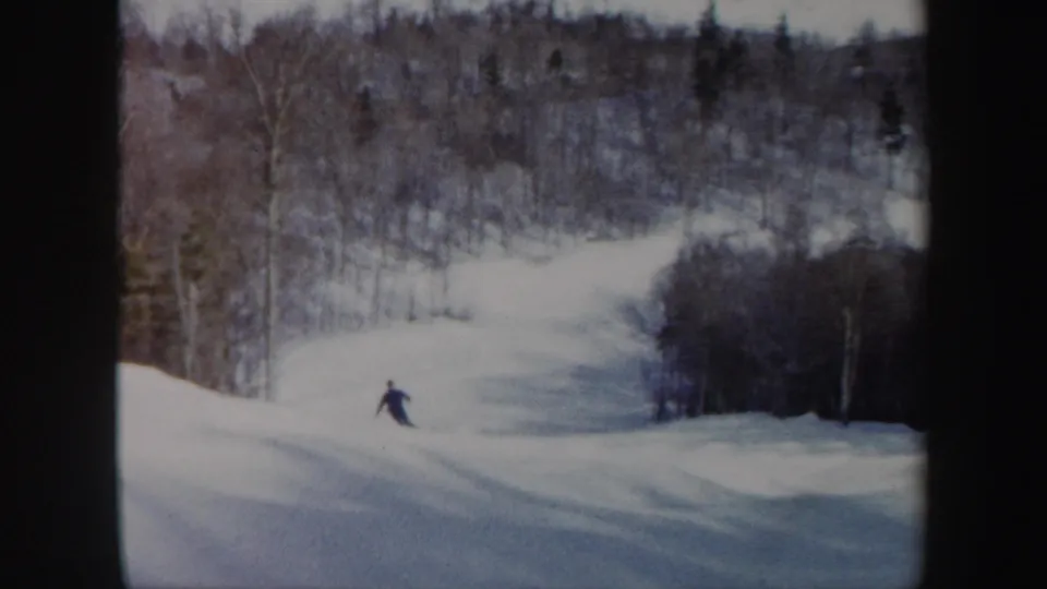 The skier continues downhill on a wooded Vermont ski slope, small in frame and surrounded by pale snow and winter trees.