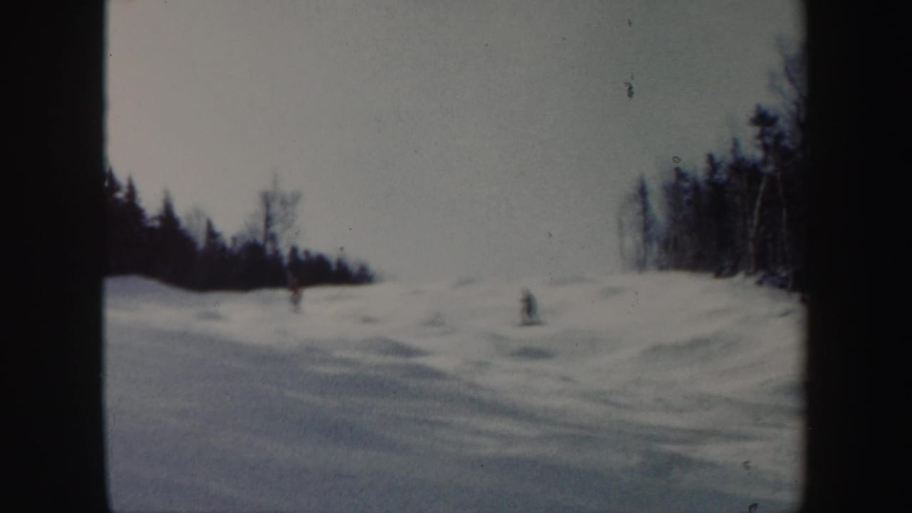 1962 Vermont 8mm frame showing small distant skiers on a broad snowy slope bordered by dark winter trees.