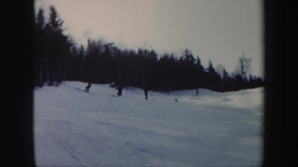 Distant skiers in dark winter clothing cross a Vermont snow slope, with dark trees and 8mm scan borders visible.