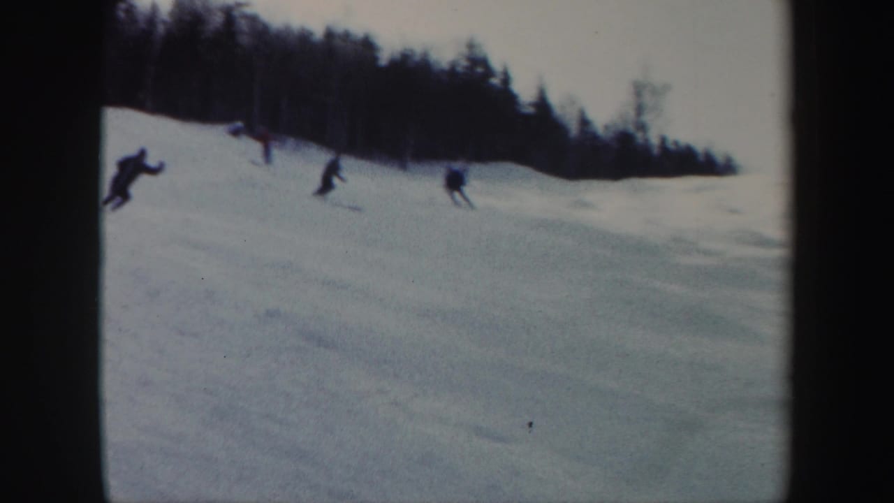 1962 Vermont home-movie frame showing multiple skiers on an open snowy slope with a dense treeline behind them.