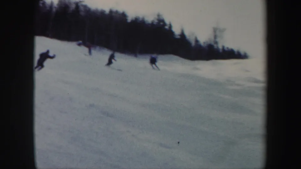 1962 Vermont home-movie frame showing multiple skiers on an open snowy slope with a dense treeline behind them.