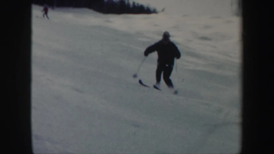 1962 Vermont 8mm frame showing a skier in dark winter clothing descending a snowy slope, with a distant skier near the upper left.