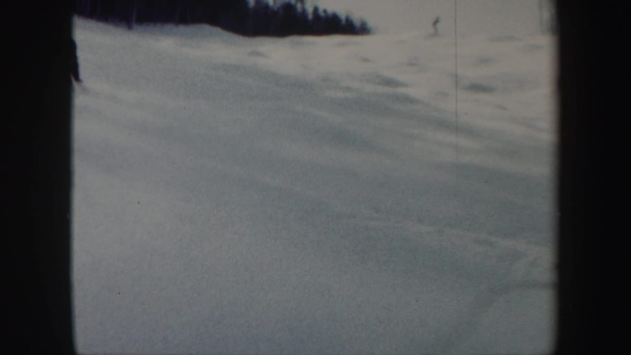 Open snowy ski slope with a distant skier near the right edge and dark trees above.