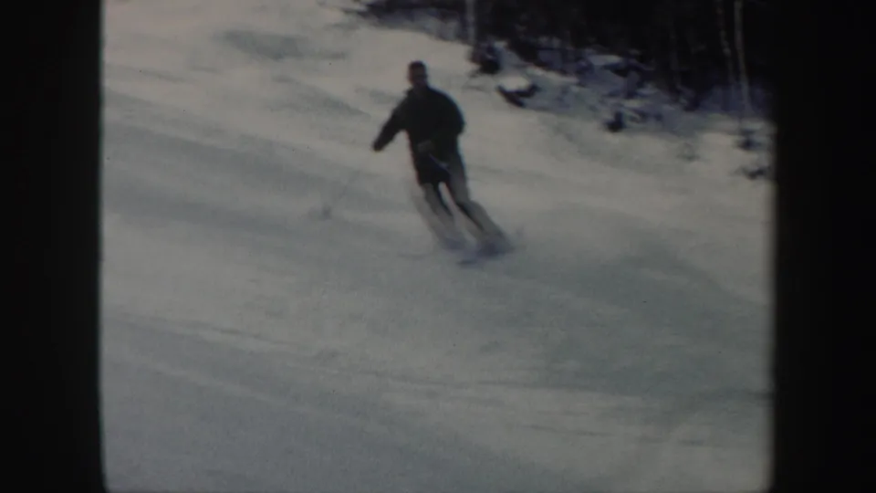 In 1962 Vermont home-movie footage, an adult man in dark ski clothes turns downhill on a snow-covered slope.