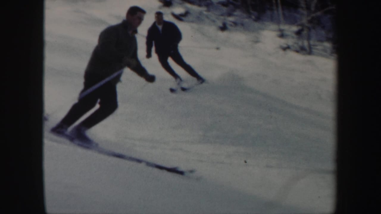 Two adult men ski downhill in Vermont, with the nearer skier leaning into a turn on the sunlit snow.