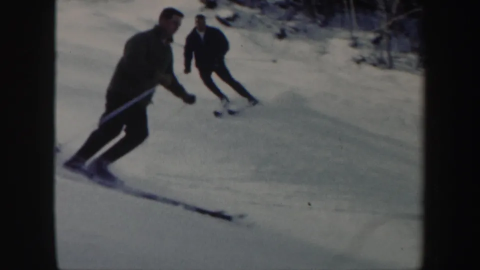 Two adult men ski downhill in Vermont, with the nearer skier leaning into a turn on the sunlit snow.