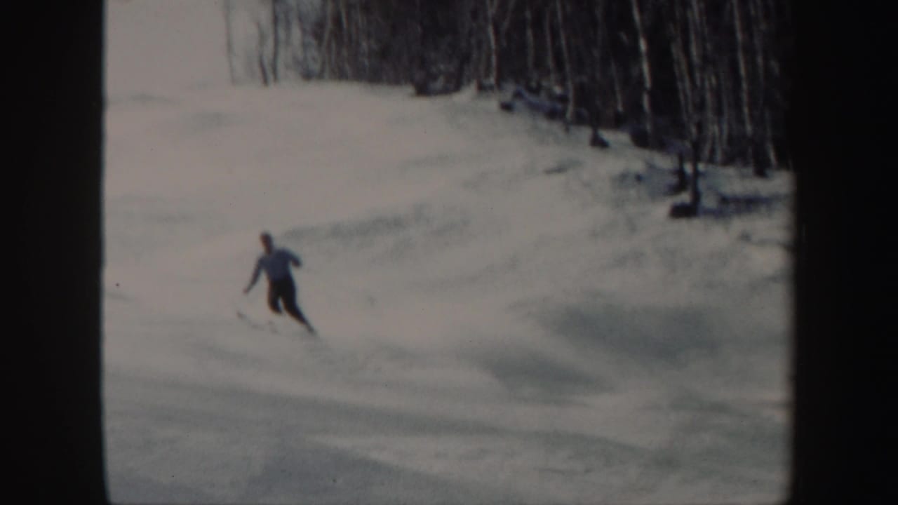 In 1962 Vermont home-movie footage, a skier in dark pants moves downhill across a snowy open slope near bare trees.