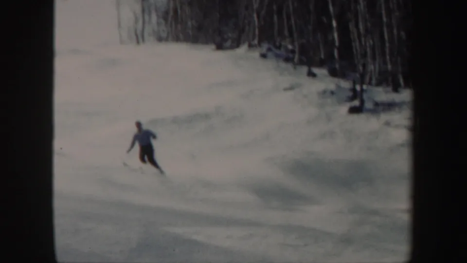 In 1962 Vermont home-movie footage, a skier in dark pants moves downhill across a snowy open slope near bare trees.