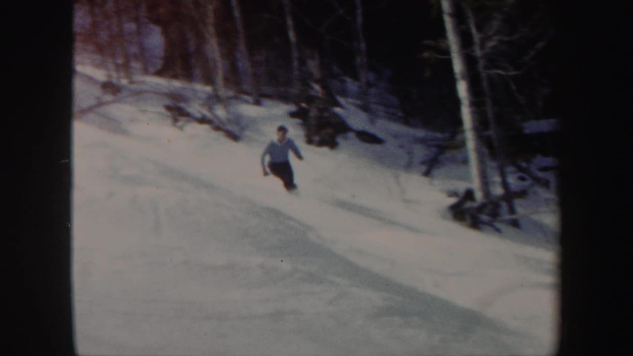 The skier continues downhill through a Vermont snowfield, with bare trees and shadows behind him.