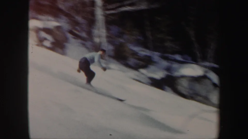 A skier in a light top and dark pants moves downhill on a snowy Vermont slope in 1962, with trees and rocks along the edge.