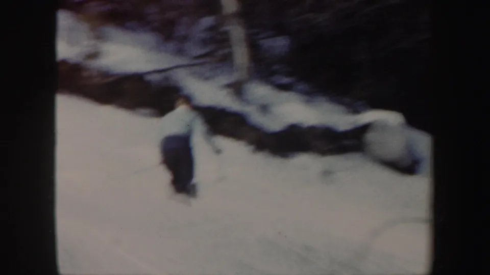 The skier moves farther down the snowy incline, partly blurred by motion, with dark trees and rounded rocks behind him.