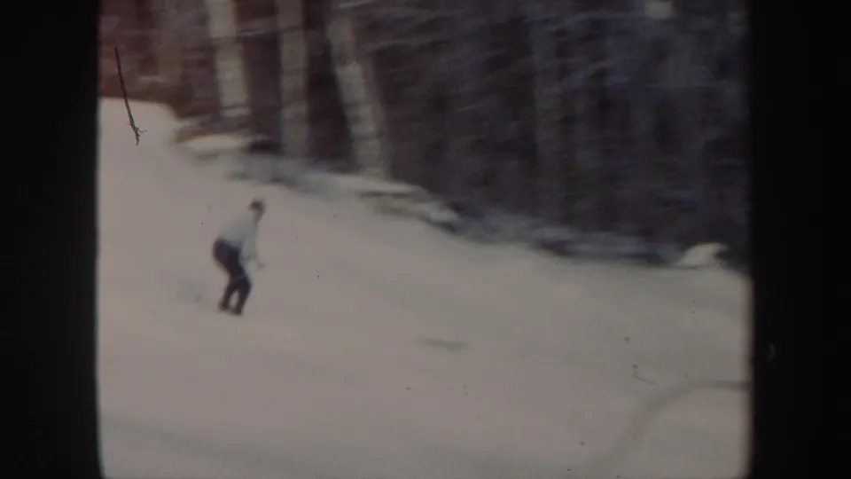 The skier appears lower on the slope, gliding across pale snow with a wooded hillside and soft home-movie blur in the background.