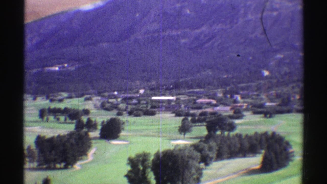 1969 Colorado home-movie frame with manicured green land, tree clusters, a small settlement, and mountain foothills.