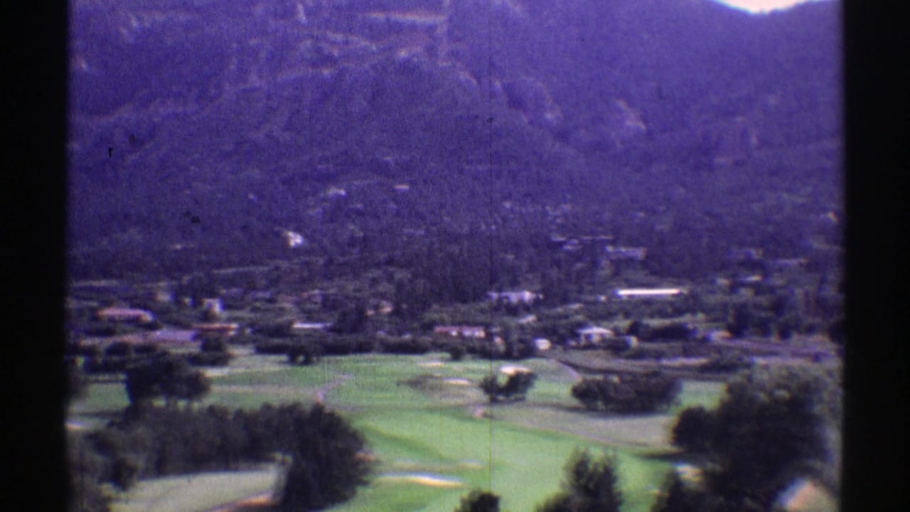1969 Colorado 8mm view of green fairways, scattered trees, low buildings, and a forested mountain rising behind them.