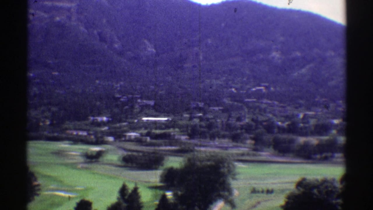 Vintage Colorado home-movie frame showing a golf-course-like green area, trees, scattered structures, and a dark mountain backdrop.