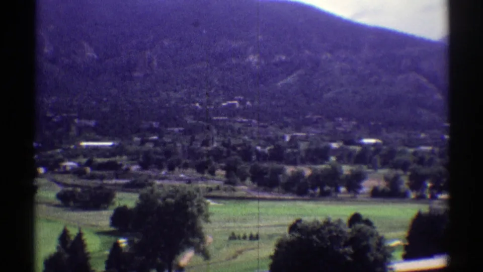 Wide 1969 Colorado landscape frame with green open ground, dark trees, small buildings, and a forested mountain across the background.