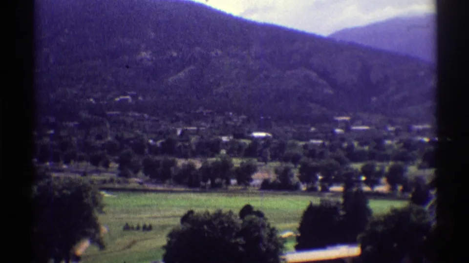 Colorado 8mm frame of a green course area, distant low buildings, dark tree lines, and a wooded mountain under clear light.
