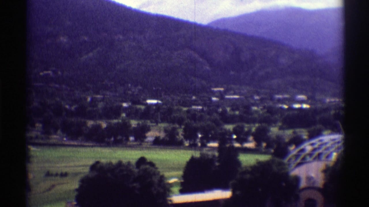 1969 Colorado home-movie frame showing a green valley village below dark mountain ridges, with an arched greenhouse structure near the foreground.