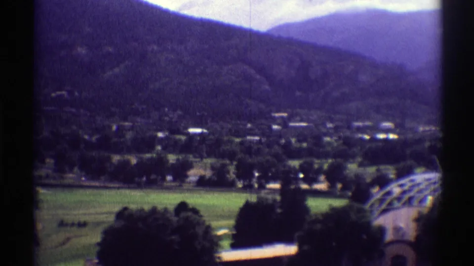 1969 Colorado home-movie frame showing a green valley village below dark mountain ridges, with an arched greenhouse structure near the foreground.