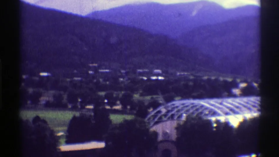 Soft 8mm frame of a 1969 Colorado valley view with scattered buildings, dark mountain slopes, and the arched greenhouse roof entering at right.