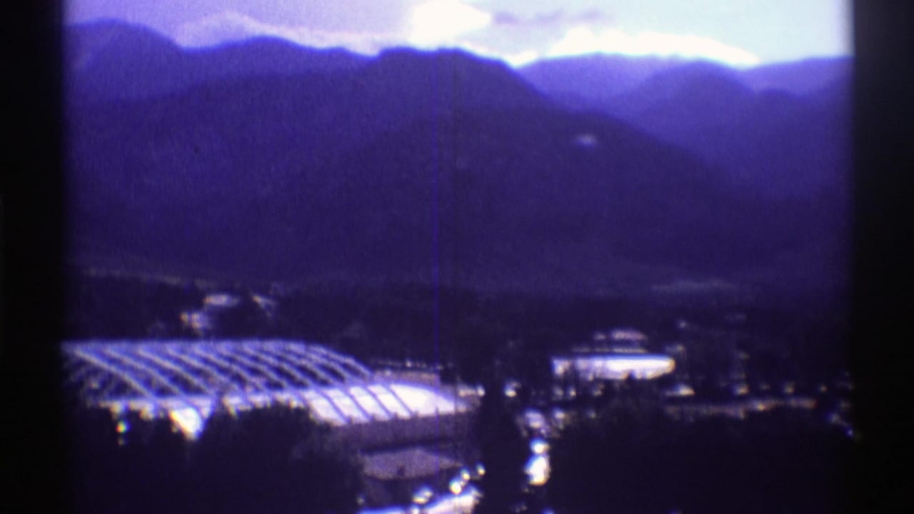 Colorado home-movie frame with a large curved roof, nearby lit structure, roads, and mountain slopes in blue-purple film color.