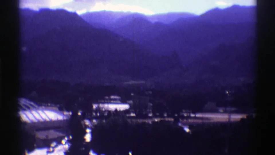 Colorado 8mm frame showing hazy mountain layers, a cropped domed roof, nearby buildings, and dark treetops from an elevated viewpoint.