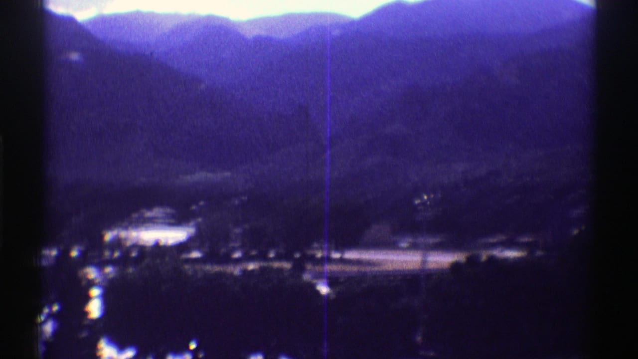 1969 Colorado 8mm frame showing a distant mountain ridge, valley trees, small settlement details, and a pale sky with purple-blue film cast.