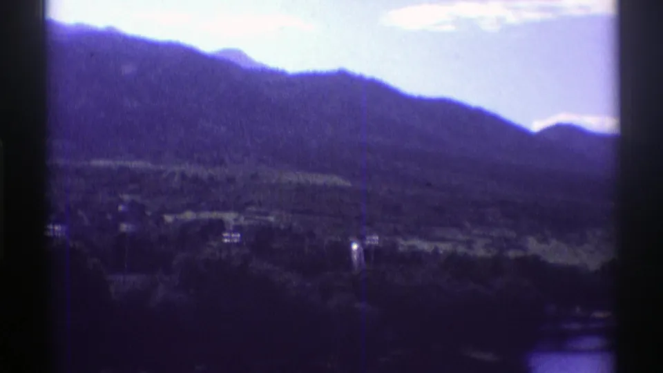 1969 Colorado home-movie frame with mountain silhouettes, scattered trees, and faint shoreline buildings.