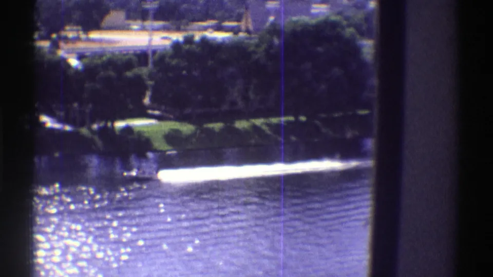 1969 Colorado 8mm frame showing a motorboat wake on a bright lake, with shoreline trees and parked cars beyond dark window framing.