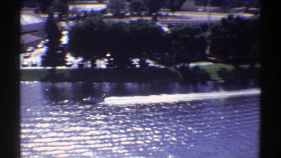 The motorboat moves across sparkling water in a 1969 Colorado home-movie view, with a tree-lined shoreline and parking area in the background.