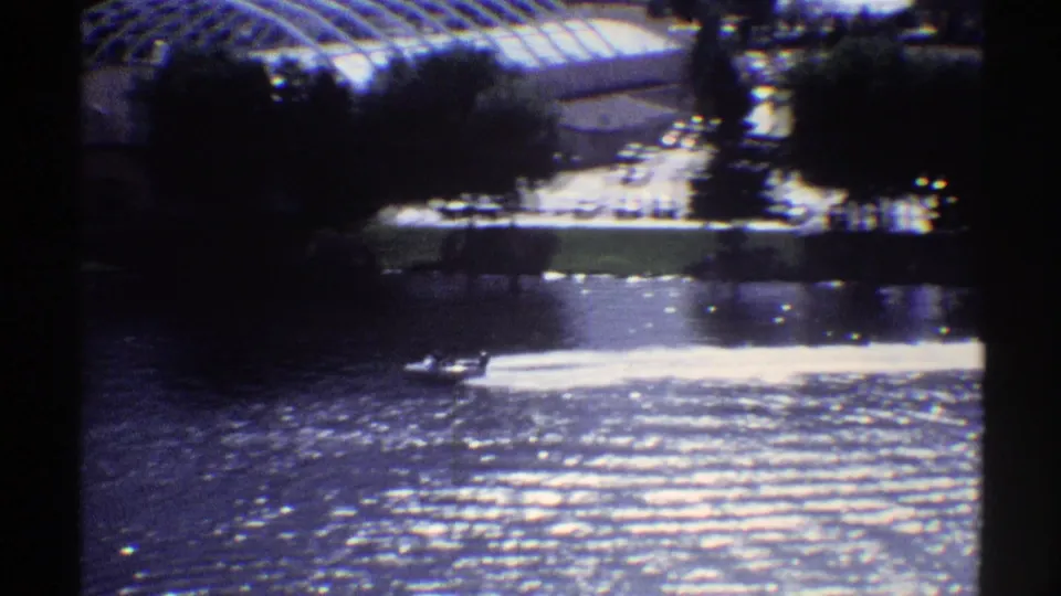 A small motorboat and bright wake pass through the lower frame, with shaded trees, a shoreline path or lawn, and a bright parking area behind.