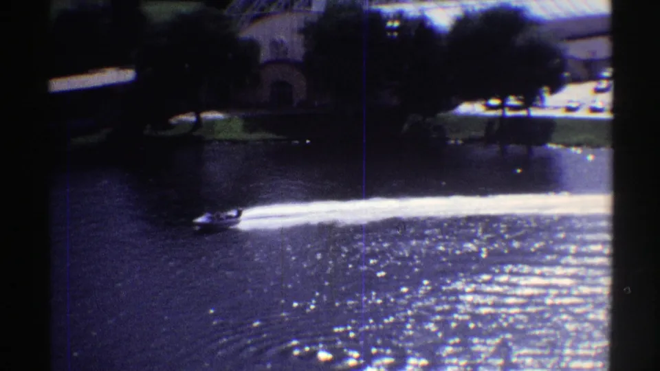 Final frame shows the boat and wake on a sun-glittered Colorado lake, with trees, shoreline lawn, and dark window framing from the filming position.