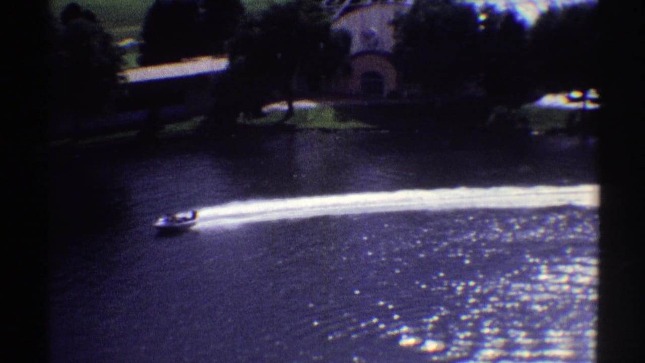 1969 Colorado 8mm frame showing a small speedboat moving across dark lake water with a bright wake and shoreline buildings behind it.