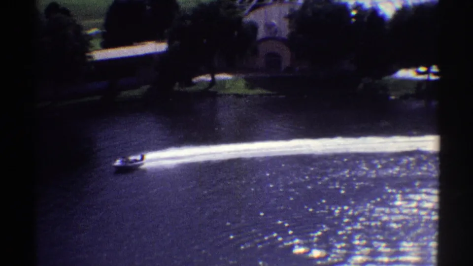 1969 Colorado 8mm frame showing a small speedboat moving across dark lake water with a bright wake and shoreline buildings behind it.
