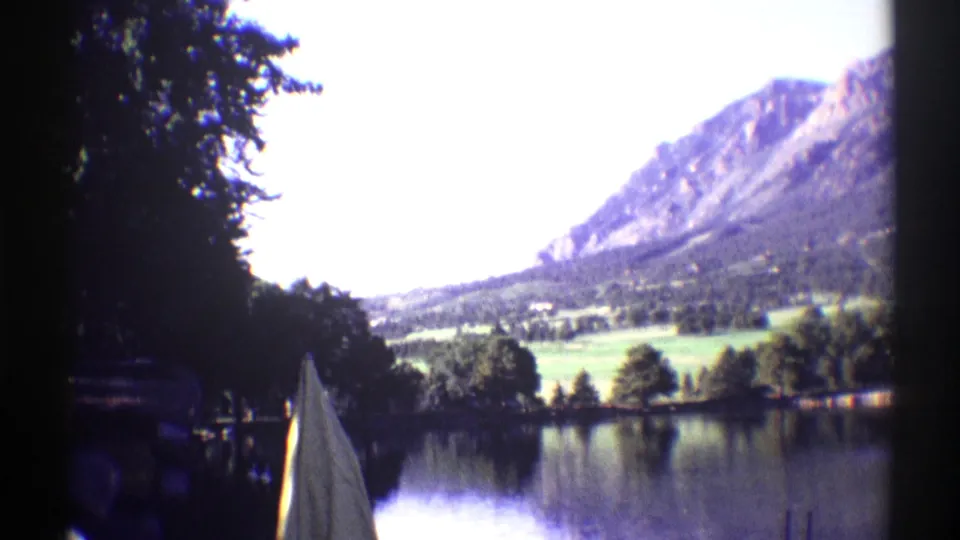 1969 Colorado 8mm frame looking across a calm lake toward trees and brown mountain slopes under a bright sky.