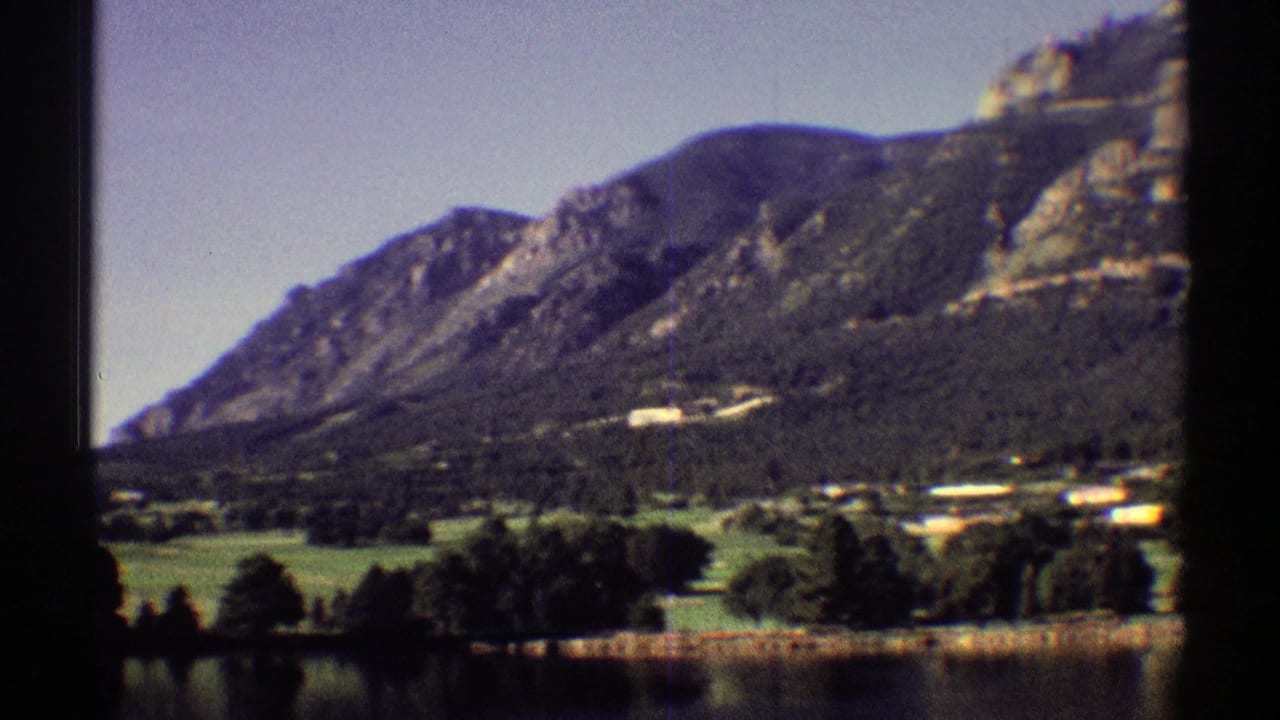 Mountain slope above green fields, trees, shoreline houses, and a calm lake.