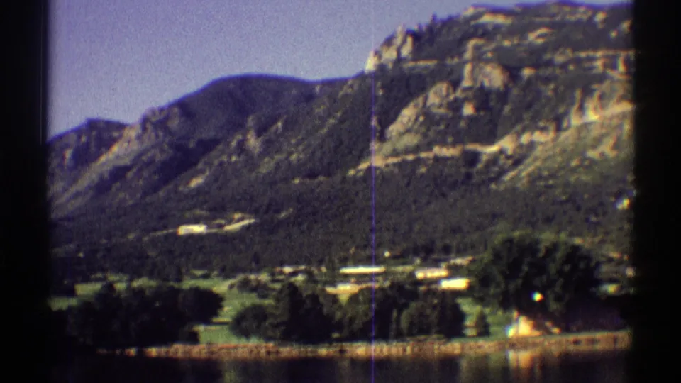 1969 Colorado home-movie view of a rocky mountain face above trees, small buildings, open grass, and lake shoreline.