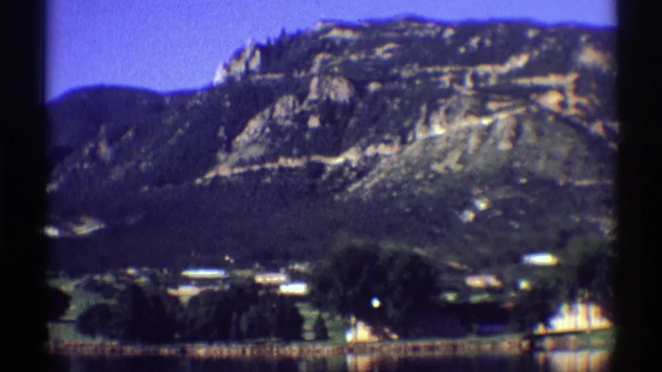 1969 Colorado 8mm frame with a rocky mountainside, dark tree cover, low buildings, grassy fields, and lake foreground.