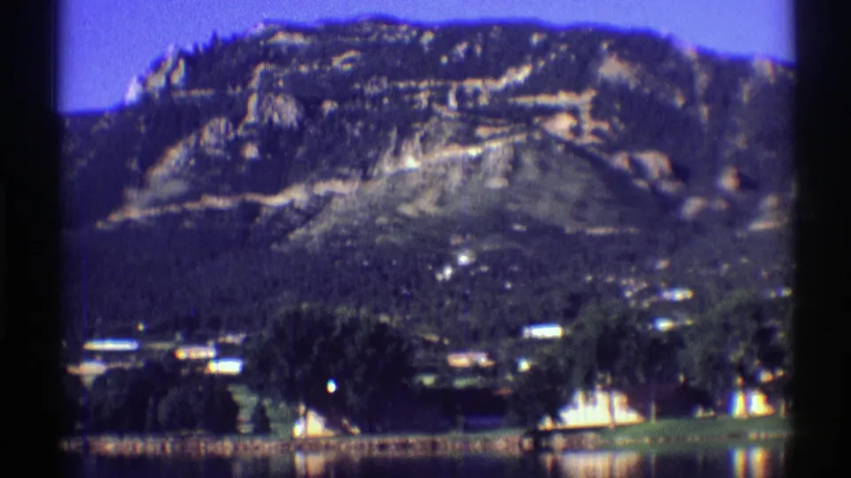 1969 Colorado frame showing the pan across a rocky mountain slope with trees, houses, grass, and still lake water below.
