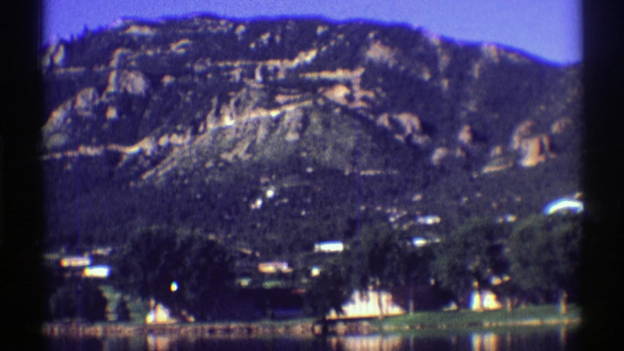 1969 Colorado 8mm frame showing a calm lake, trees, small shoreline structures, and rugged mountain slopes in the distance.